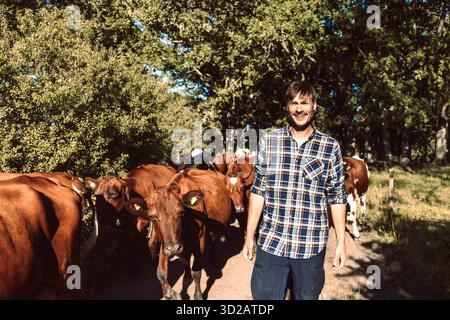 Lächelnder Vieharbeiter, der mit einer Kuhherde auf Gehwegen auf dem Bauernhof läuft Stockfoto