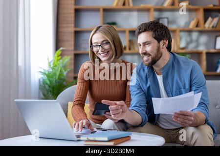 Lächelndes Paar, das auf einem Sofa sitzt, ihre Haushaltsausgaben und Investitionen plant, Zahlen mit einem Laptop und Taschenrechner berechnet, Rechnungen organisiert Stockfoto