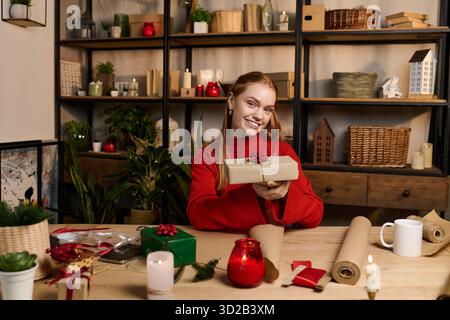 Die junge Frau in einem roten Pullover hüllt die Geschenke zu Hause und schafft eine warme und festliche Atmosphäre. Stockfoto