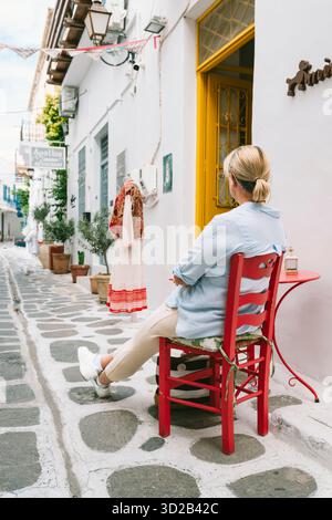 Eine blonde Frau sitzt auf einem roten Stuhl in einer Gasse in der Altstadt von Parikia, Paros, Griechenland. Stockfoto