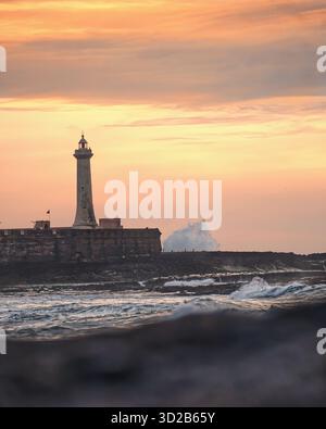 Eine ruhige Küstenszene mit einem hohen Leuchtturm, der auf einem Steinpier unter einem warmen, pastellfarbenen Sonnenuntergang steht. Die Wellen des Ozeans schlagen sanft auf Stockfoto