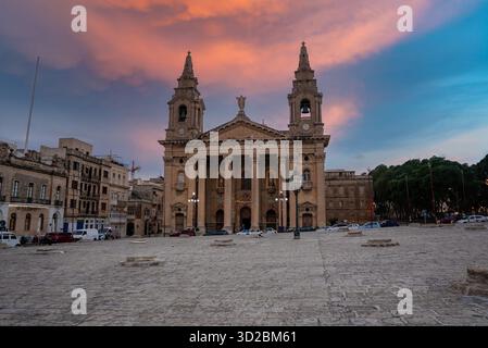 Eine barocke Kirche mit zwei Türmen und einem Säulengang blickt bei Sonnenuntergang auf eine piazza aus Stein in Valletta, Malta. Warmes Licht leuchtet auf Kalkstein, Autos parken in der Nähe, La Stockfoto
