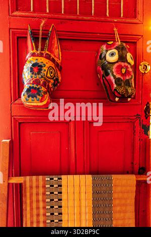 Merida, Yucatan, Mexiko, zwei Masken hängen an einer roten Tür. Die Masken sind farbenfroh und haben ein einzigartiges Design. Die Tür ist rot und hat einen Holzrahmen Stockfoto