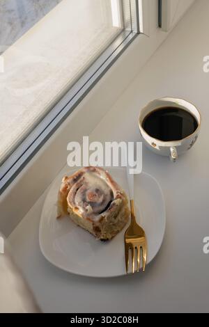 Eine gemütliche Szene mit einer frisch gebackenen Zimtrolle auf einem weißen Teller mit einer goldenen Gabel und einer Tasse schwarzen Kaffee auf einer Fensterbank. Stockfoto