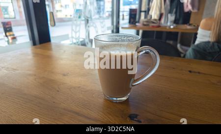 Heißer Kaffee in Glasbecher auf Holztisch in der Nähe eines hellen Fensters im Café, moderne Atmosphäre ideal für Lifestyle und Entspannungskonzepte. Stockfoto