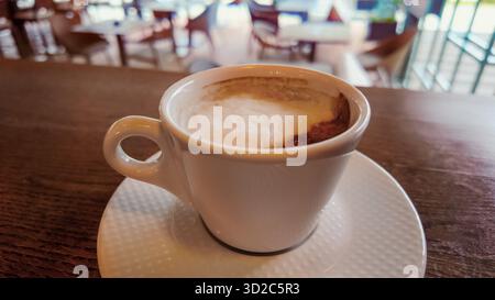 Nahaufnahme einer heißen Kaffeetasse mit Schaum auf Holztheke, verschwommener Café-Hintergrund, der Wärme, Aroma und Entspannung betont Stockfoto