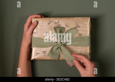Die Kinderhände halten ein Weihnachtsgeschenk, lösen das Band und packen ein Weihnachtsgeschenk aus, das in goldfarbenes Papier gewickelt ist und mit einem grünen Band auf einem grünen olivfarbenen Rücken gebunden ist Stockfoto