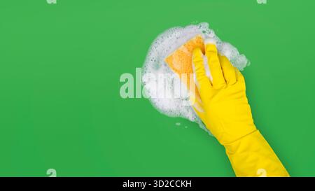 Hand in Yellow Handschuh hält Schwamm und wischt seifigen Schaum auf grünem Hintergrund ab Stockfoto
