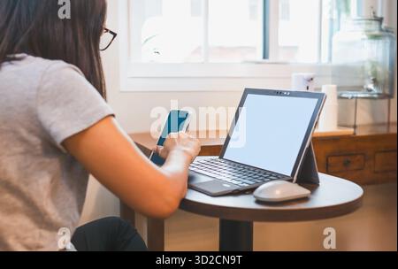 Die Frau benutzt das Handy, während sie im Café mit dem Laptop arbeitet. Stockfoto