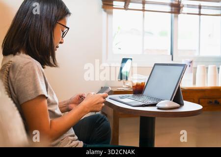Die Frau benutzt das Handy, während sie im Café mit dem Laptop arbeitet. Stockfoto