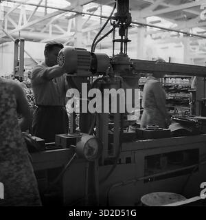 Archiv Schwarzweißfoto aus den 1970er Jahren in Sloviansk, Ukrainische SSR. Es zeigt einen männlichen Arbeiter, der eine große Industriemaschine im Armature-Isolator Plant (AIZ) bedient. Er steht an seinem Arbeitsplatz und konzentriert sich darauf, ein Bauteil, möglicherweise einen Elektromotor, an der komplexen Maschine anzupassen. Im Vordergrund ist ein Manometer zu sehen. Der Hintergrund zeigt den Innenraum der Werkstatt. Dieses Bild ist ein klassisches Beispiel für einen sowjetischen Industriearbeiter, der in der Produktion tätig ist Stockfoto