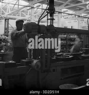 Archiv Schwarzweißfoto aus den 1970er Jahren in Sloviansk, Ukrainische SSR. Es zeigt einen männlichen Arbeiter, der eine große Industriemaschine im Armature-Isolator Plant (AIZ) bedient. Er steht an seinem Arbeitsplatz und konzentriert sich darauf, ein Bauteil, möglicherweise einen Elektromotor, an der komplexen Maschine anzupassen. Im Vordergrund ist ein Manometer zu sehen. Der Hintergrund zeigt den Innenraum der Werkstatt. Dieses Bild ist ein klassisches Beispiel für einen sowjetischen Industriearbeiter, der in der Produktion tätig ist Stockfoto