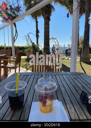 Eine erste-Person-Aussicht von einem Holztisch in einem Café im Freien, mit einem Obstbecher und einem Getränk im Vordergrund. Im Hintergrund stehen Palmen und Meer Stockfoto