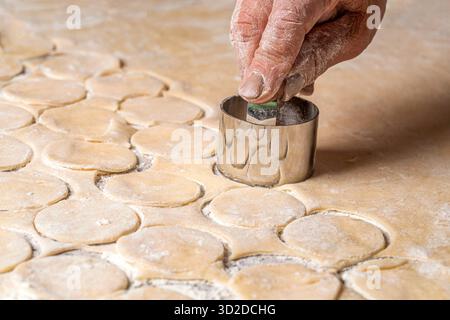 Zubereitung des Teigs zum Backen, ein großes Stück des Teigs liegt auf dem Tisch und der Koch schneidet kleine Kreise mit eiserner Form aus. Stockfoto