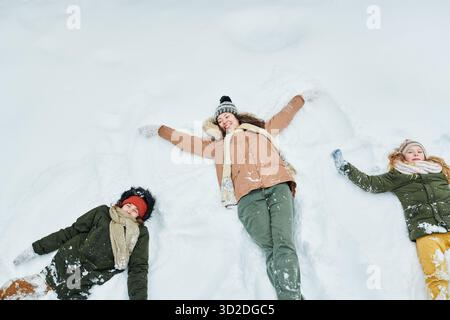 Kaukasische Frau liegt im Schnee mit zwei kaukasischen Kindern, die Schneeengel basteln, alle lächeln und aufschauen, Winterkleidung sichtbar, Winteraktivität im Freien, Familienverbindungsmoment Stockfoto