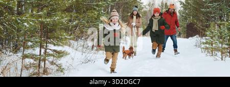 Zwei kaukasische Kinder, die mit einem kleinen braunen Hund durch einen verschneiten Waldweg laufen, während Kaukasierin und Kaukasier hinter sich her folgen, alle in Winterkleidung und genießen Outdoor-Aktivitäten Stockfoto
