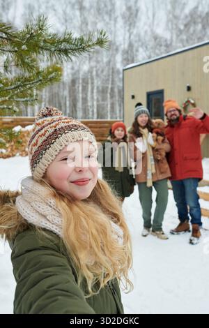 Porträt eines kaukasischen Mädchens, das draußen in verschneiten Landschaften vor der Kamera lächelt, mit einer multiethnischen Familie, einschließlich Mutter, Vater, Bruder und Hund, die im Hintergrund stehen Stockfoto