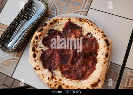 Frische Pizza mit dünnen Scheiben Räucherfleisch, serviert im Freien mit Besteck und Gewürzen. Italienisches Café im Freien, Blick von oben. Stockfoto