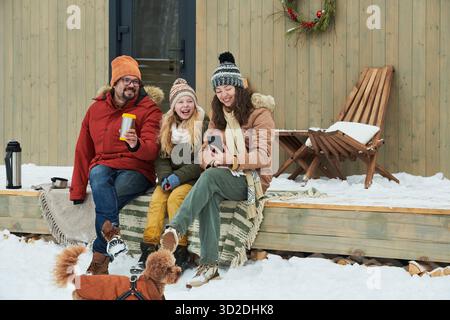 Kaukasische Familie, die in Winterkleidung auf einer hölzernen Veranda sitzt, lächelt und auf das Smartphone blickt, während ein kleiner Hund im Schnee spielt Stockfoto