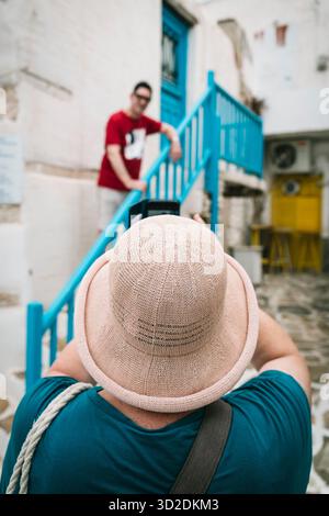 Eine Frau mit Strohhut fotografiert mit ihrem Handy einen Mann auf einer Treppe in den Gassen der Altstadt von Antiparos, Kykladen, Griechenland. Stockfoto