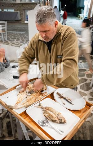 Ein Mann schneidet gegrillten Fisch zum Abendessen an einem Tisch in einem Restaurant an der Hafenpromenade, Aliki, Paros, Griechenland Stockfoto