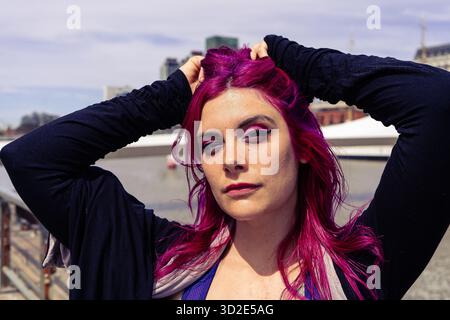 Frau mit auffälligen magentafarbenen Haaren in legeren Kleidern, die sich selbstbewusst im Freien vor einem urbanen Hintergrund posiert Stockfoto