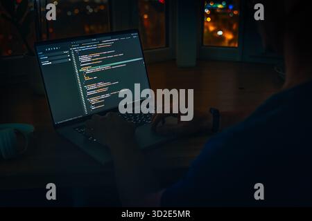 Die Hände des Programmierers programmieren auf einem Laptop im Dunkeln mit Blick auf die Lichter der Nachtstadt. Das Konzept eines Entwicklers oder Hackers. Stockfoto