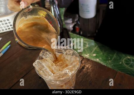 Erfrischendes Eistee-Getränk, das im Café aus dem Glas in eine Tasse Eis gefüllt wird. Zufriedenstellendes Milchtee-Getränk, das für den Gast auf einem Holztisch zubereitet wird Stockfoto