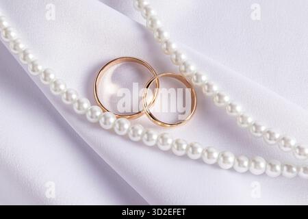 Zwei elegante goldene Eheringe liegen auf einer großen Falte aus weißem Satin zwischen Perlensträngen. Draufsicht. Das Konzept der Hochzeit. Postkarte Stockfoto