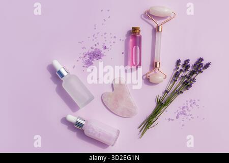 Ein Set von Kosmetikprodukten in Glasflaschen, ein Massagegerät und ein Rosenquarzschaber, frischer Lavendelzweig. Flache Lagen. Draufsicht. Home Spa Care Stockfoto