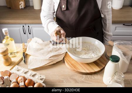 Die Hand einer Frau rührt das Mehl mit einem Schneebesen in einer Schüssel mit den Zutaten für den Teig. Butter, Eier, Milch, eine Dose Mehl auf dem Küchentisch Stockfoto