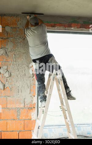 Zwei Bauarbeiter verputzen und glätten die Decke während der Innenrenovierung in einem Gebäude mit Ziegelwänden. Professionelle Bauarbeiter arbeiten Stockfoto
