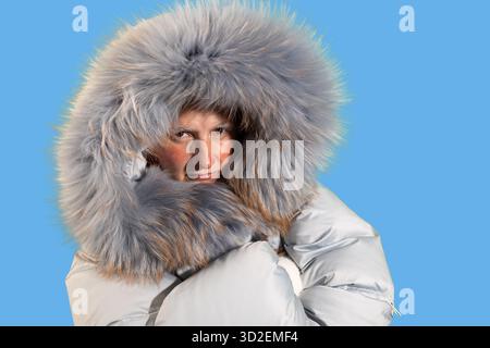 Eine Frau mit Frost an den Augenbrauen und der Nase trägt einen geschwollenen Mantel mit einer großen Pelzhaube. Im Winter zeigt sie auf hellblauem Hintergrund auf die rechte Seite des Zuschauers. Stockfoto