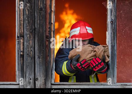 Ein Feuerwehrmann rettet ein Baby aus einem brennenden Gebäude. Der Feuerwehrmann ist in Aktion und rettet das Kind aus der Notsituation. Dieses Bild kann verwendet werden, um Themen von Tapferkeit, Rettungseinsätzen und Rettungsdiensten zu veranschaulichen. || Modell freigegeben Stockfoto