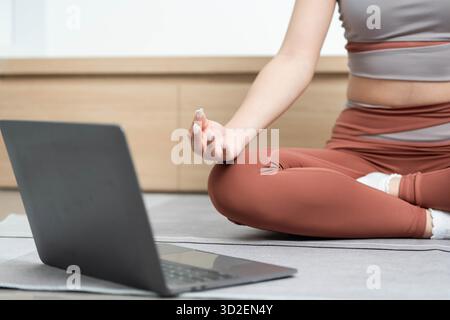 Eine junge Frau meditiert zu Hause und nutzt einen Laptop für geführte Yoga-Sitzungen, um ihre Wellness-Reise zu verbessern. || Modell freigegeben Stockfoto