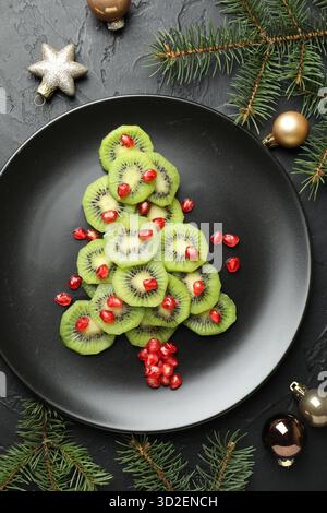 Weihnachtsbaum aus Kiwi, Granatapfelkernen und festlicher Dekoration auf schwarzem Tisch, flache Ladefläche Stockfoto