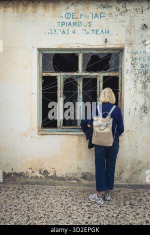 Eine Frau schaut durch ein kaputtes Fenster in ein verlassenes Gebäude in der Altstadt von Marpissa, Paros, Griechenland. Stockfoto