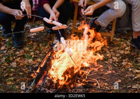 Freunde treffen sich am Lagerfeuer, rösten Würstchen auf Stöcken und genießen den Herbstabend im Freien. Gemütliches Picknick mit Leuten, die Essen über hellen Flammen kochen Stockfoto
