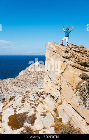 Ein Mann steht in einer Siegespose auf einem felsigen Gipfel vor dem Leuchtturm im Paros Park Nature Reserve, Griechenland. Stockfoto