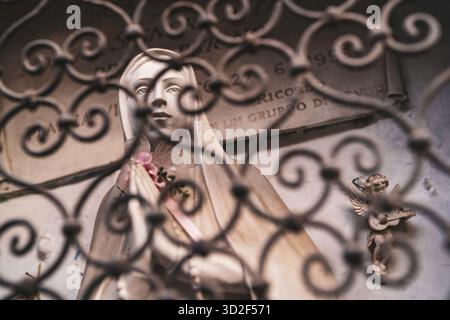 marienstatue mit gefalteten Händen und einer kleinen rosafarbenen Rose, durch ein kunstvoll verziertes schmiedeeisernes Tor gesehen, mit einem Engel, der eine Mandoline in weichem f spielt Stockfoto