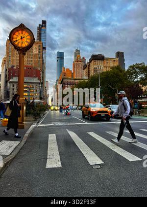 Abendlicher Blick auf die Fifth Avenue in der Nähe des Flatiron Building, New York City, mit Fußgängern, die die Straße überqueren, gelben Taxis und einer Vintage-Uhr. Stockfoto