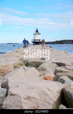 Die Leute laufen entlang des felsigen Stegs, der zum Spring Point Ledge Lighthouse führt, mit herbstlichen Bäumen, die über die Casco Bay sichtbar sind Stockfoto