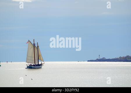 Ein Schoner segelt unter ruhigem Himmel in Portland Harbor in Richtung Horizont, wobei der entfernte Leuchtturm Portland Head Light entlang der Küste sichtbar ist. Stockfoto