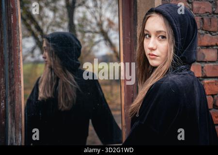 Teenager in einem schwarzen Hoodie steht an einem alten Fenster und sieht ihr Spiegelbild an. Selbstreflexion, Einsamkeit oder emotionales Geschichtenerzählen Stockfoto