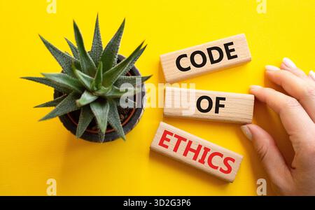 Symbol für Ethikkodex. Konzeptworte Ethik-Kodex auf Holzblöcken. Geschäftsmann Hand. Schöner gelber Hintergrund mit saftiger Pflanze. Business und Stockfoto