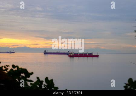 Ein Blick auf die Küste von verankerten Frachtschiffen bei Sonnenuntergang, mit warmen Reflexen über dem Wasser und Berglandschaften unter einem leuchtenden Himmel Stockfoto