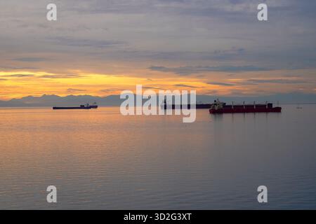 Verankerte Frachtschiffe ruhen auf glasklarem Wasser unter einem glühenden Sonnenuntergang, mit fernen Bergen und weichen Wolken, die eine friedliche Küstenatmosphäre bilden. Stockfoto