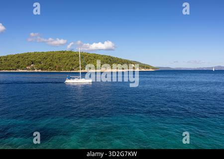 Eine ruhige Meereslandschaft mit einem einsamen Segelboot, das auf tiefblauem Wasser gleitet, einem felsigen Vordergrund und einer grünen Insel in der Ferne unter einem hellen, klaren Sch Stockfoto