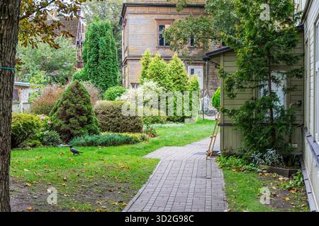 Ein gepflasterter Weg führt durch einen üppigen, angelegten Garten im Hinterhof mit verschiedenen Evergreens und einem historischen, verwitterten gelben Holzhaus im Hinterhof Stockfoto