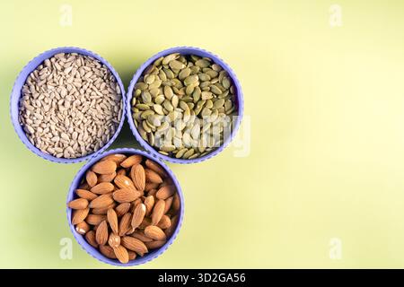 Drei violette Schüsseln mit verschiedenen Nüssen und Samen auf grünem Hintergrund. Die Schalen sind dreieckig angeordnet Stockfoto
